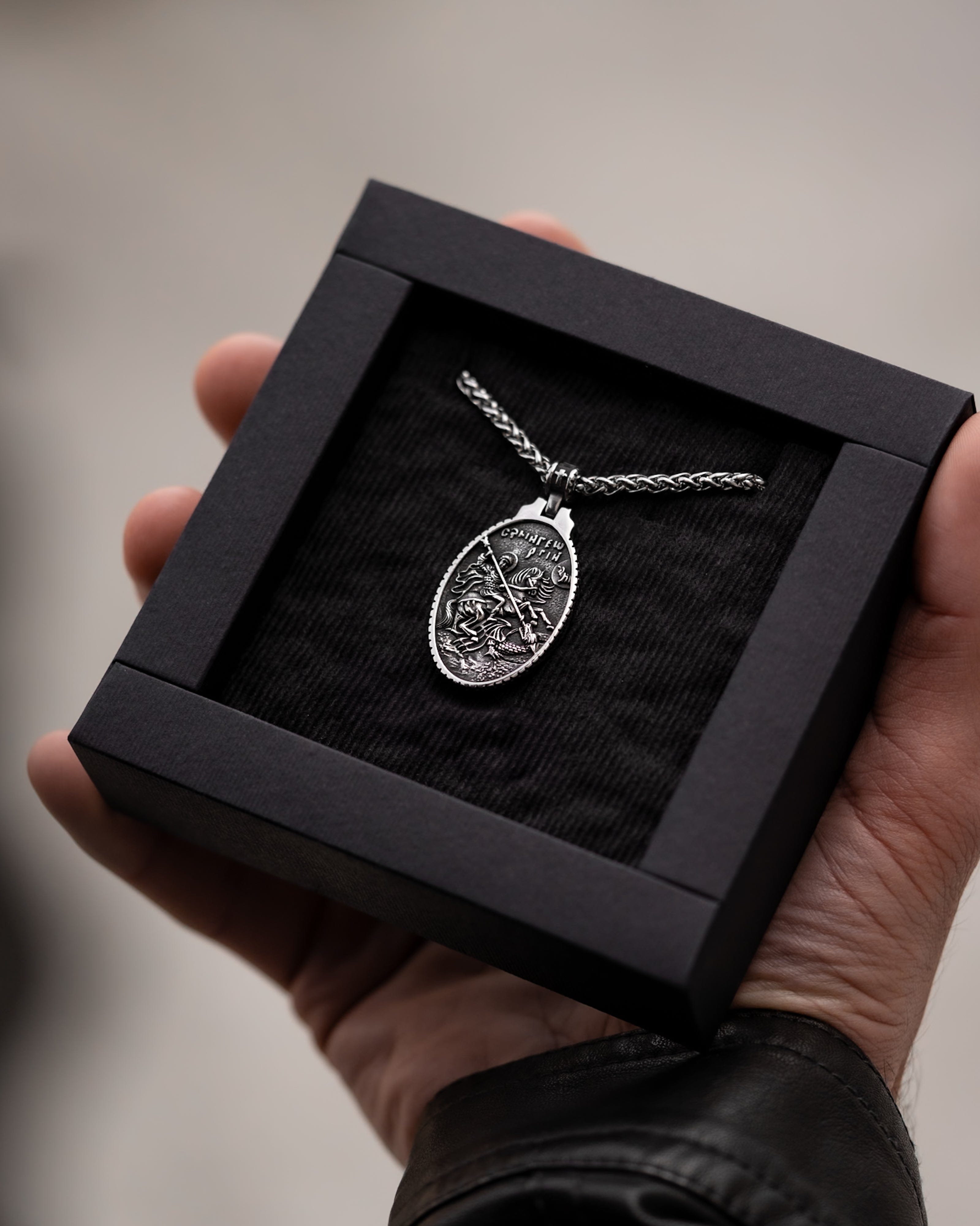 Muskarac drzi i pokazuje kutiju sa muskom ogrlicom SV. Georgije Silver, Privezak i lanac HIRUSKI CELIK, srebrna boja, Najbolja cena i prodaja u Srbiji, model 25
